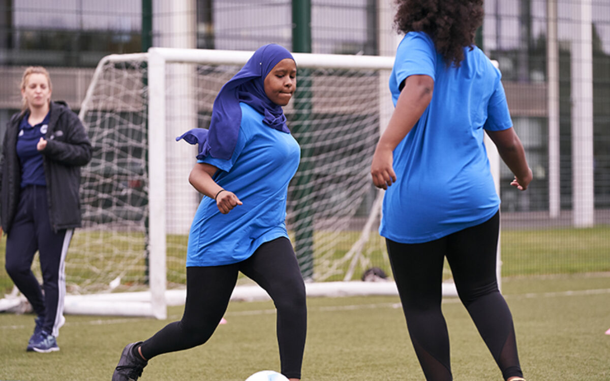 Teen girl in hijab kicking a ball about to be tackled by another teen girl